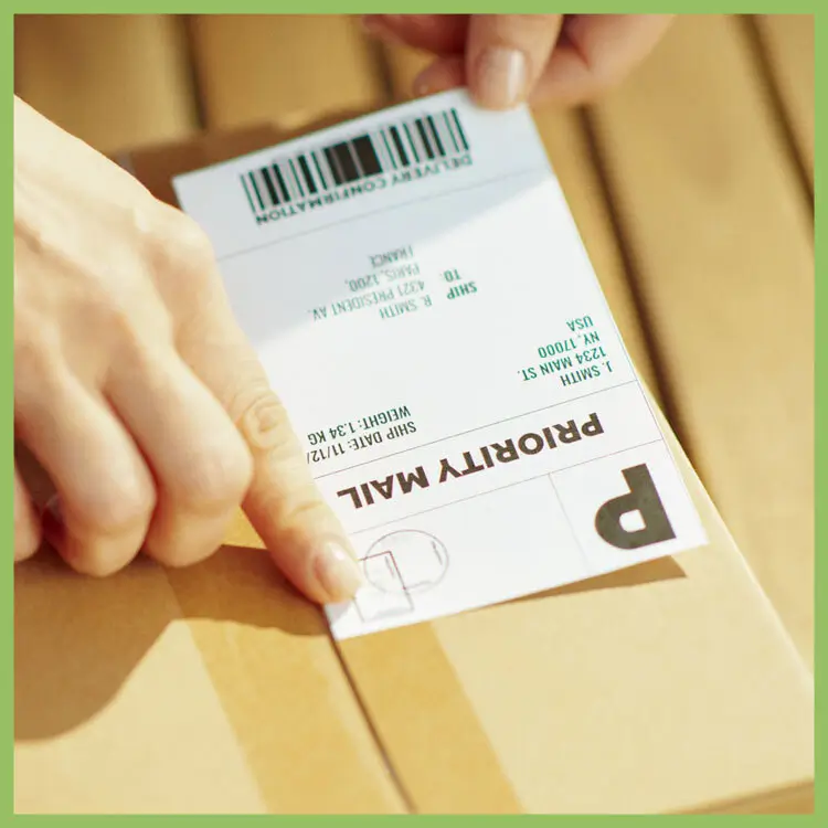 Hands applying a shipping label with barcode and Priority Mail marking onto a cardboard box
