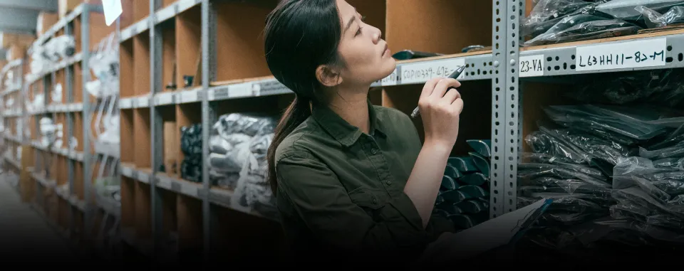 Warehouse staff member conducting manual stocktake using a clipboard while inspecting inventory items on shelves, surrounded by neatly organised stock.